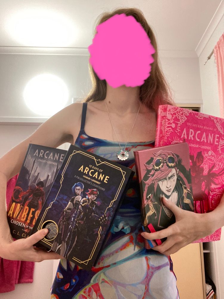 Someone with their face scribbled out wearing the Arcane Blackmilk anomaly dress and the RockLove jayvik anomaly cog, holding Ambessa: Chosen of the Wolf, the 12 Days of Arcane Advent Calander, the sculpted Vi notebook, and the making of Arcane art book