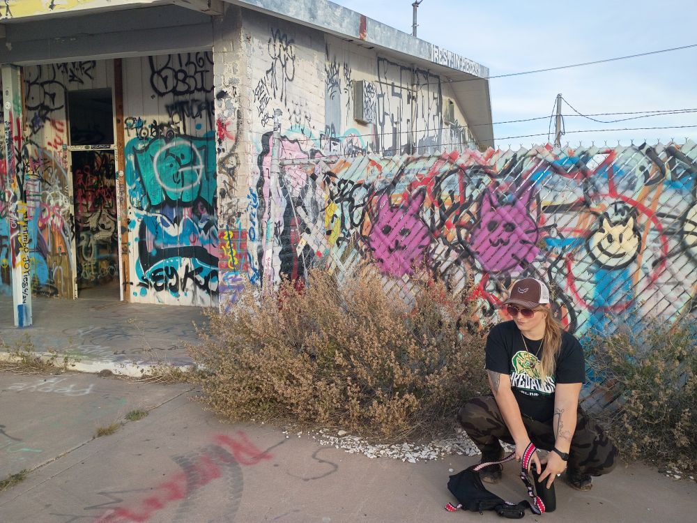 Buff white woman crouching below cat graffiti at old gas station above the Apache death cave 