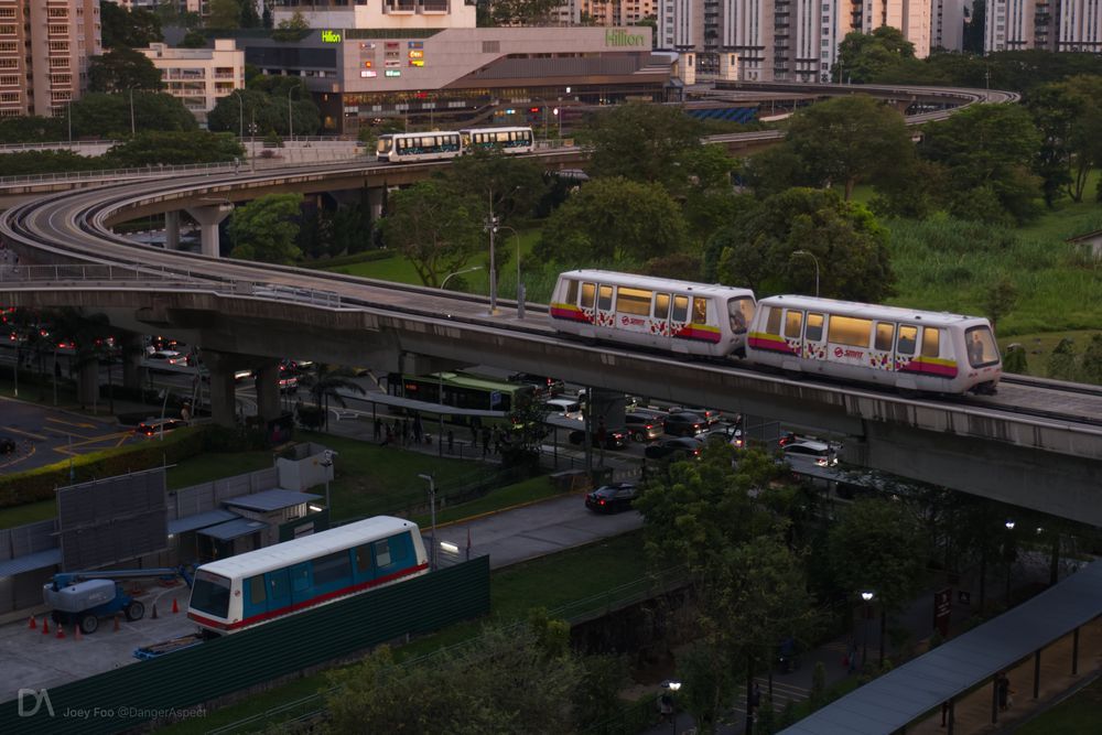 An elevated view showing a fenced site office in the bottom corner, with LRT viaducts snake overhead. In the site office, a first-generation LRT vehicle sits in a corner. Overhead, a coupled pair of second-generation vehicles are traveling past, and in the distance, a new coupled pair of third-generation vehicles is approaching in the opposite direction.