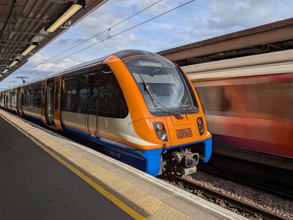 A London Overground Class 710 sitting at a platform while a London Underground S stock rushes past behind. 