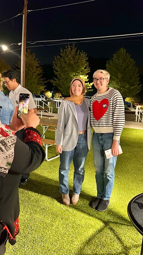 White woman wearing a tan and navy hijab, jeans, and tan shoes standing to the left of a white person with blond hair wearing black and white striped shirt with a big red heart and jeans standing on grass
