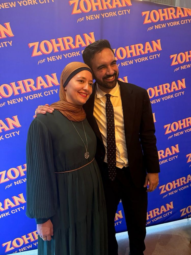 Back ground with Zohran for New York City logo on blue. Ruwa and Zohran are standing in front of it. Ruwa is on the left wearing a tan and black scarf with green dress. Zohran is on the right wearing a black suit, white shirt, and black tie with white polka dots
