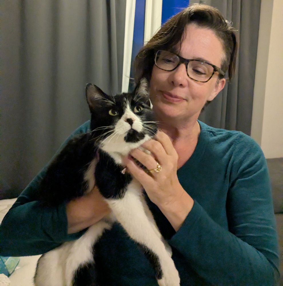 Photo of me (middle-aged white woman with dark hair and glasses) holding a black and white cat in our Airbnb apartment in Grundafjordur, Iceland. Vanda is enjoying some chin skritchies. 