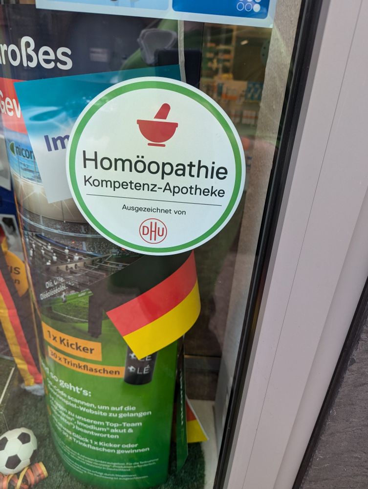Eine Apotheke hat außen einen Sticker mit "Homöopathie Kompetenz Apotheke - Ausgezeichnet von DHU" an ihrem Schaufenster angebracht
