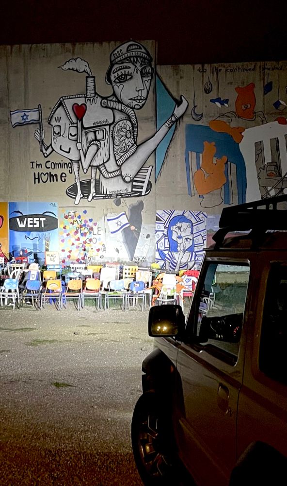 Ein Auto strahlt eine Betonwand mit Graffiti an. Das größte zeigt eine s/w gezeichnete Person, die einen Rucksack trägt, der wie ein Haus mit Armen aussieht und der in einer Hand eine Israelfahne und in der anderen ein rotes Herz trägt. Darunter steht „I‘m coming Home“