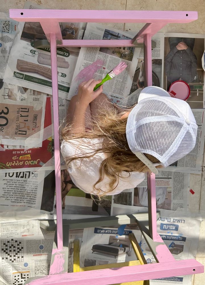 Ein kleines Mädchen sitzt in einem Tischgestell auf Zeitungspapier mit einem Malerpinsel in der Hand. Die Farbe am Pinsel ist Rosa und das Tischgestell teilweise auch. 
