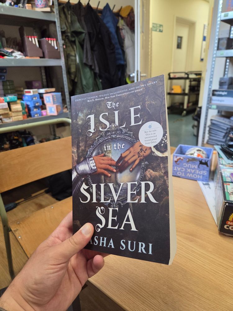 A photo of a hand holding up a large size paperback book, The Isle In The Silver Sea by Tasha Suri, with the front cover facing the camera and the right- hand edge visible to give an impression of the thickness of the book. The cover shows the hands of two South Asian women touching, one wearing a gauntlet, over a circular opening through which a castle on the sea is visible. The cover also has a small white circle with the text "Not for sale. This is an uncorrected bound proof from Orbit."