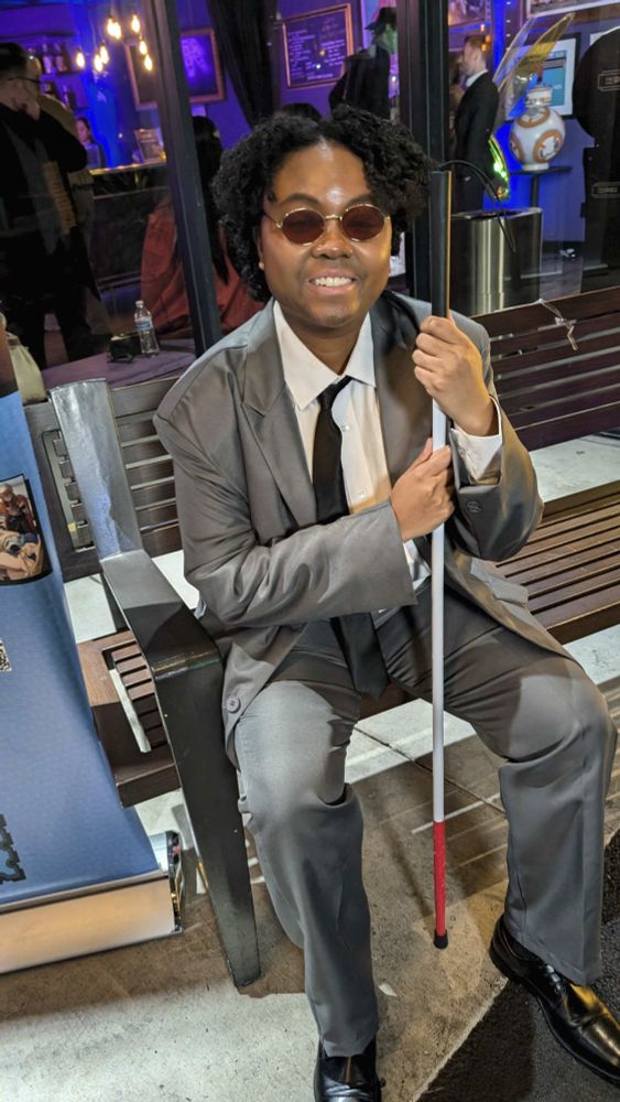 patchouliies dressed as Matthew Murdock from Marvel's Daredevil sitting on a bench holding a prop blind cane.