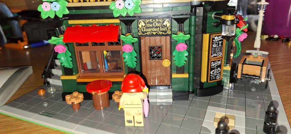 A close-up of the front of the pub. A lady wearing a rain hat and pink umbrella is looking at the pub. The walls are green, in front are a table made out of a barrel and two stools. On the corner is a menu. 