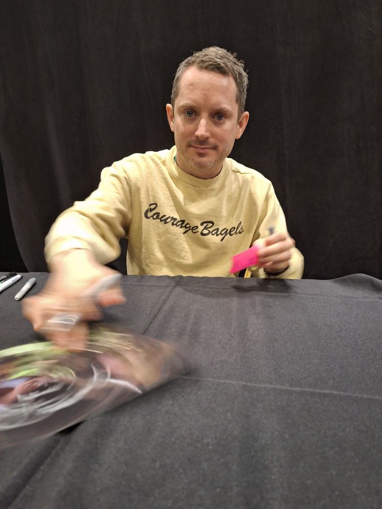 Elijah Wood spinning a signed picture while sitting at a booth table.