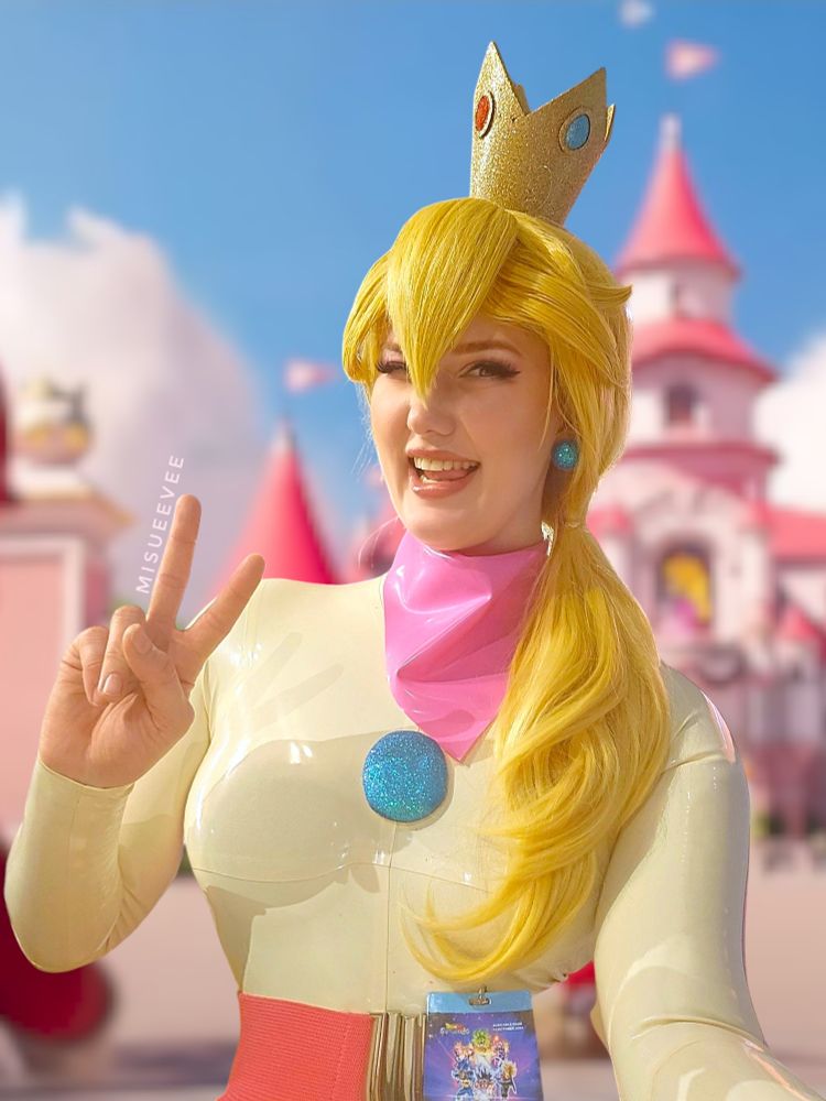 Misueevee cosplaying as Princess Peach from the Mario franchise, wearing a cream-colored latex outfit with a pink latex scarf, a large blue glittery gem accessory, and a golden crown. She has long blonde hair styled in a side ponytail and is posing with a cheerful smile, making a peace sign gesture. The background features a whimsical castle scene reminiscent of the Mario universe.