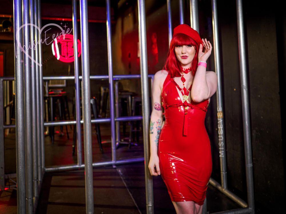 Alt Text: Eevee stands confidently in a vibrant red latex dress paired with a matching beret and necklace. Her bright red hair and bold look are accentuated by her playful pose, set against a backdrop of steel bars and dim club lighting. Neon accents in the background add a vibrant and edgy atmosphere. 