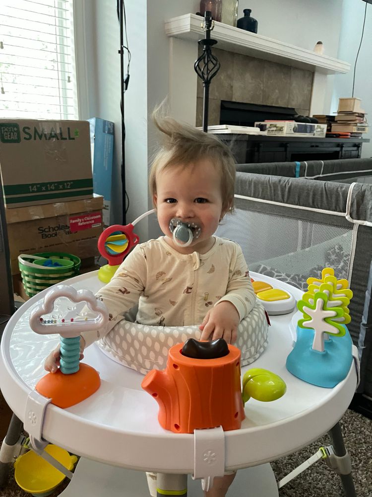A baby smiling in a standup play seat with an enormous cowlick 