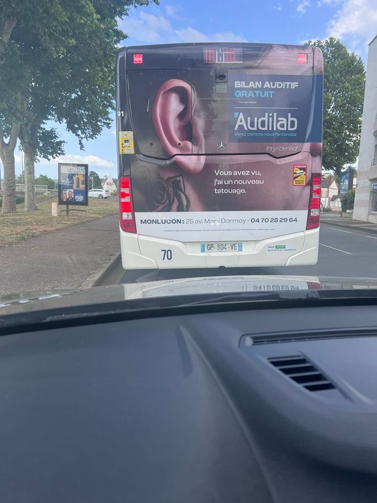 Une publicité Audika sur un bus avec une faute d'orthographe "Monlucon" au lieu de Montluçon. 
Environnement de ville avec le bus en second plan et notre voiture au premier plan.