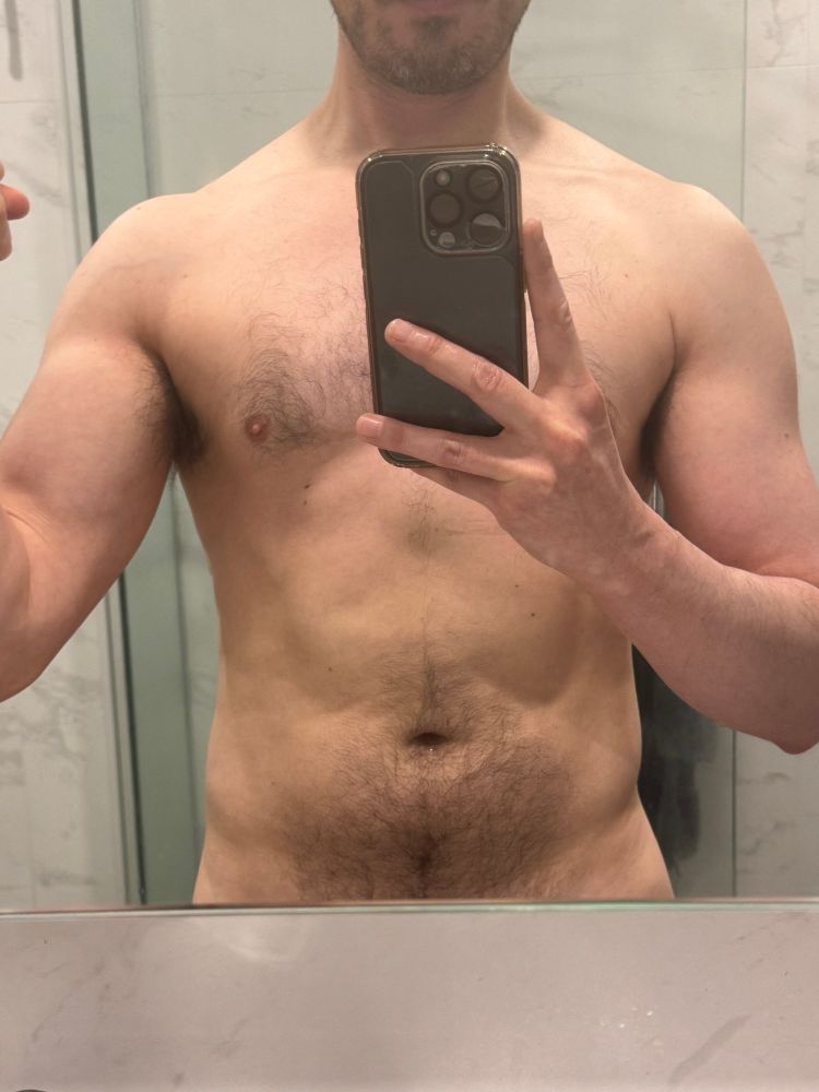 Headless torso, adifferentjon in a nude post-gym mirror selfie at home, cropped portrait showing off arms day progress with a flexed bicep, short salt and pepper beard, light chest hair, and dense stomach hair