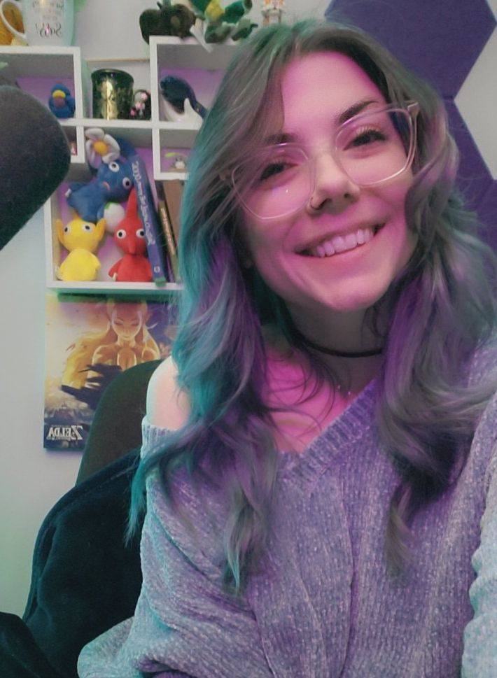 Cecilia has medium length dusty blue/gray curly hair. She is wearing glasses with clear frames and has a black hoop nose ring. She is wearing a light blue sweater off her shoulder with a black choker. Behind her is a geometric square shelf with a variety of trinkets, such as Pikmin plushes and an ocarina. This is her streaming background