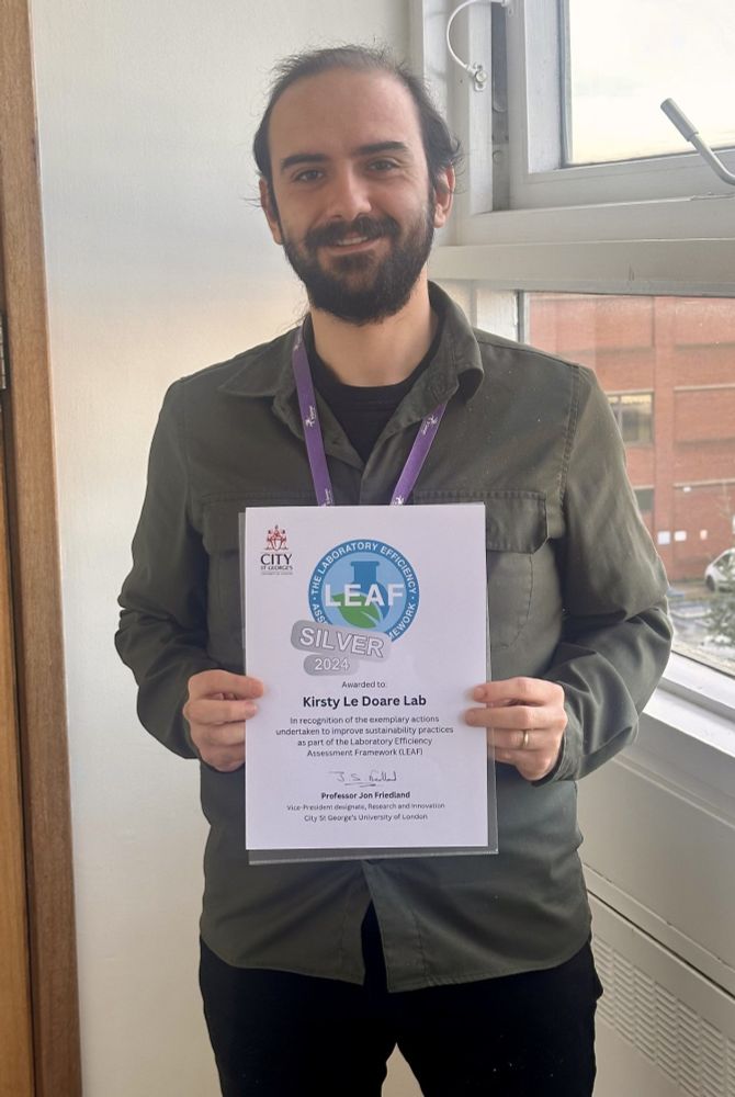 Cagil Kocana, a research scientist at the CNPI, stands in front of a window smiling while holding an award that reads "LEAF Silver 2024. Presented to Kirsty Le Doare Lab".