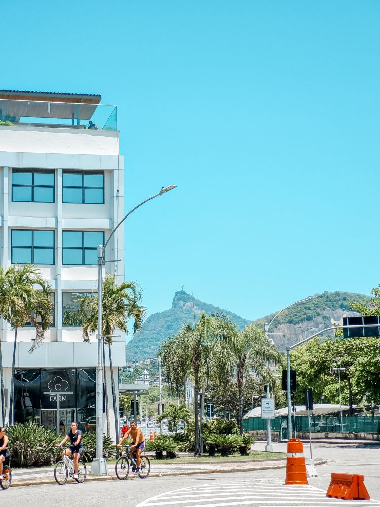 Vista do aeroporto Santos Dumont, no Rio de Janeiro. Bastante arborizado, ciclistas passeiam pela rua, o céu está azul e ao fundo o monumento do Cristo Redentor no topo do Corcovado.