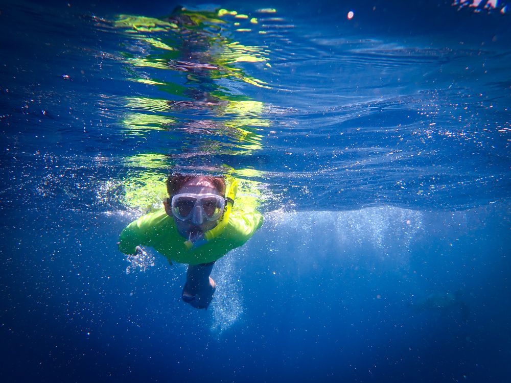 A young boy in a neon yellow shirt and a snorkel mask swims toward the camera.