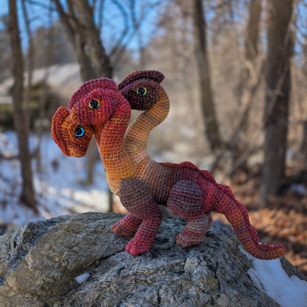 A crochet hydra in red, orange, and pink variegated yarn. It has three necks and crested heads. It stands on a rock in front of a forest.