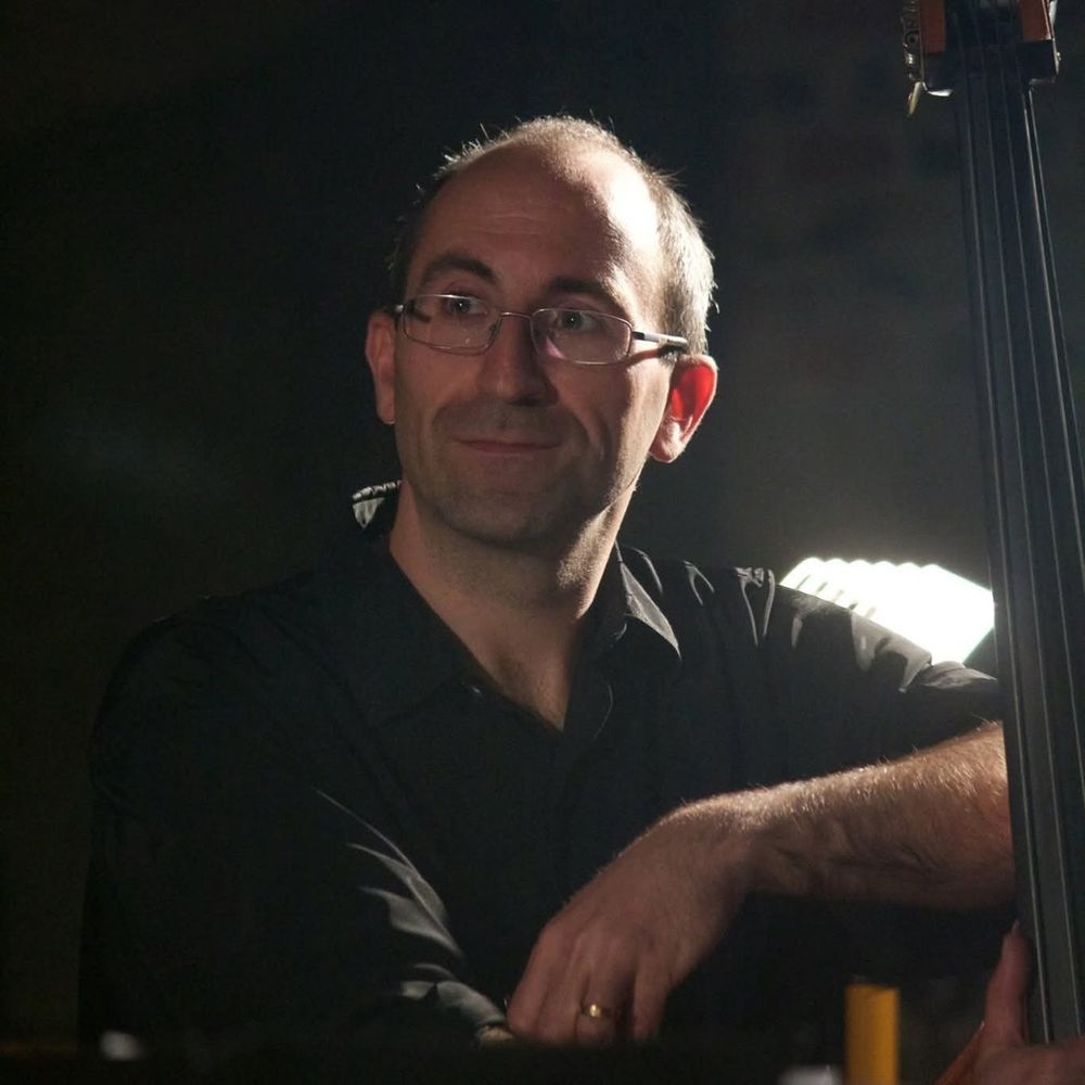 Un músic amb ulleres i camisa negra recolzat en el seu contrabaix, amb un mig somriure embadalit, envoltat d'una llum tènue 