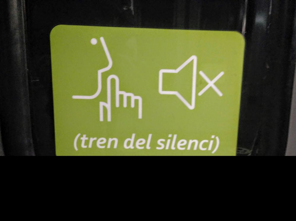 Un cartell de color verd amb un pictograma d'algú aixecant un dit davant la boca, un altre pictograma amb un altaveu silenciat, i a sota un text que anuncia, entre parèntesi, "tren del silenci"