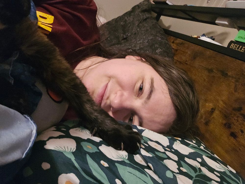 A white woman lays on a pillow, with a tortoiseshell cat's paw stretched out in front of her face, claws digging into the pillow's green and white pillowcase. 