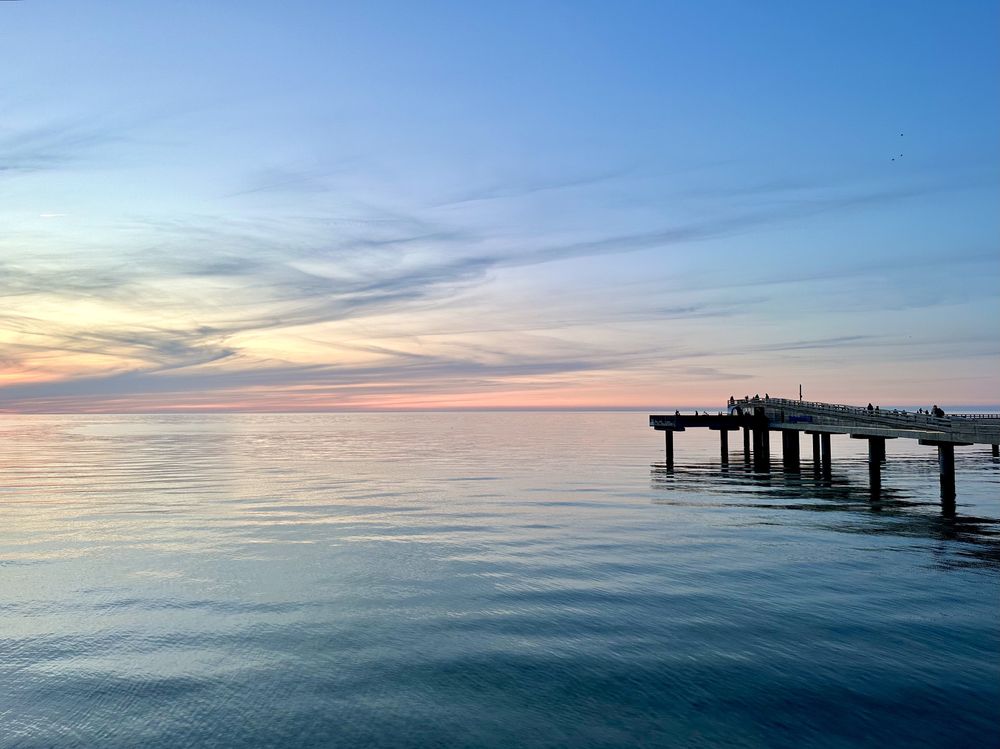 Eine friedliche Szene, die einen hölzernen Pier zeigt, der sich bei Sonnenuntergang über das ruhige Wasser erstreckt, mit sanften Pastellfarben am Himmel und einigen silhouettierten Figuren auf dem Pier.