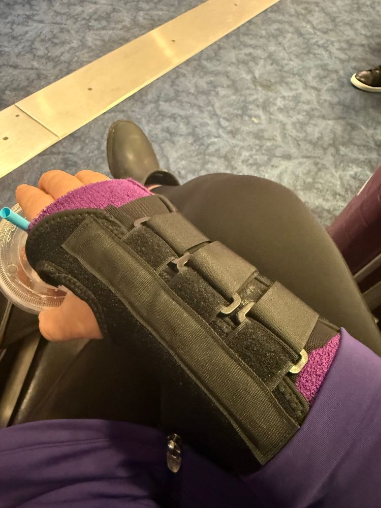 stepha is sitting at the airport gate, holding a dunkies iced tea with her surgery hand “because it counts as physical therapy” and is wearing a purple terry cloth glove liner under a black velcro wrist brace 