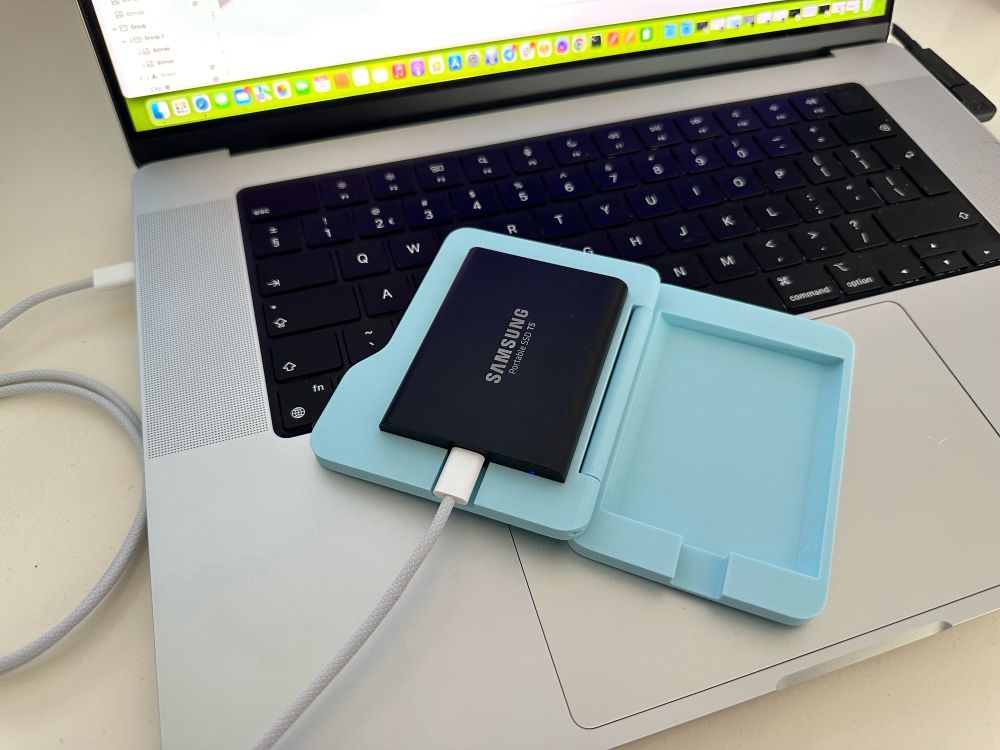 3D printed, blue Samsung SSD case in the shape of the macOS system folders. Folded open. Photographed on the top of a MacBoom Pro keyboard. Connected to the laptop with an USB-C cable.