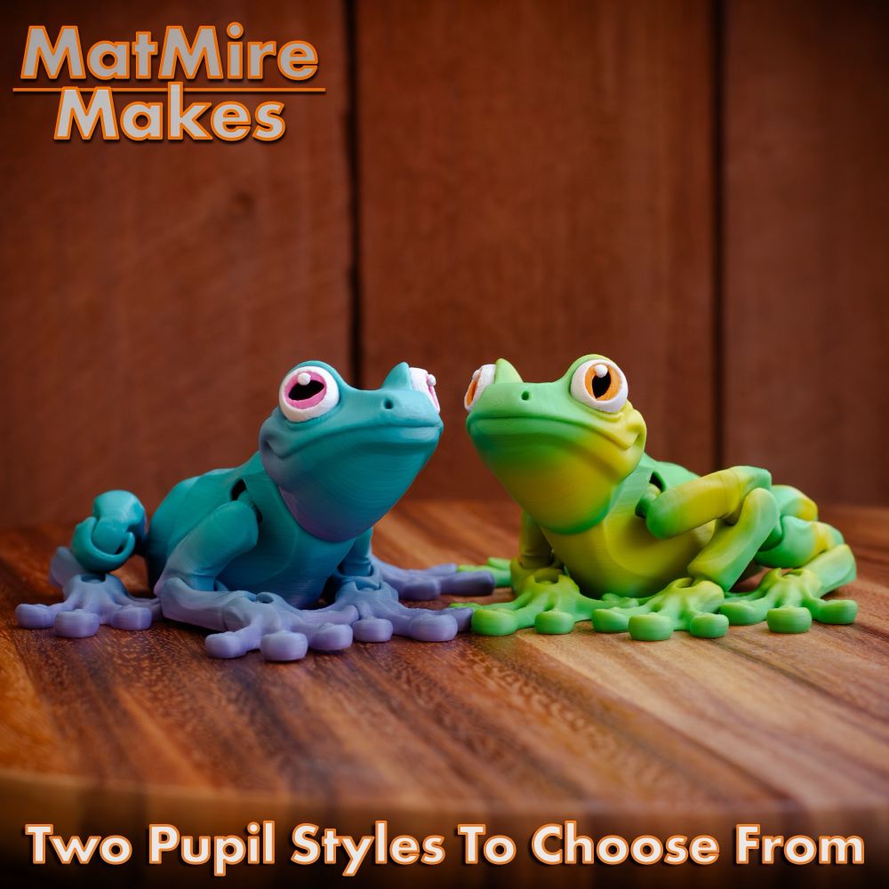 Green and purple-blue 3D printed frogs. Sitting on a wooden table with plank wall in the background.
