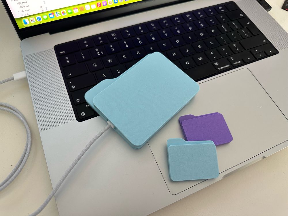 3D printed, blue Samsung SSD case in the shape of the macOS system folders. Photographed on the top of a MacBoom Pro keyboard. Connected to the laptop with an USB-C cable.