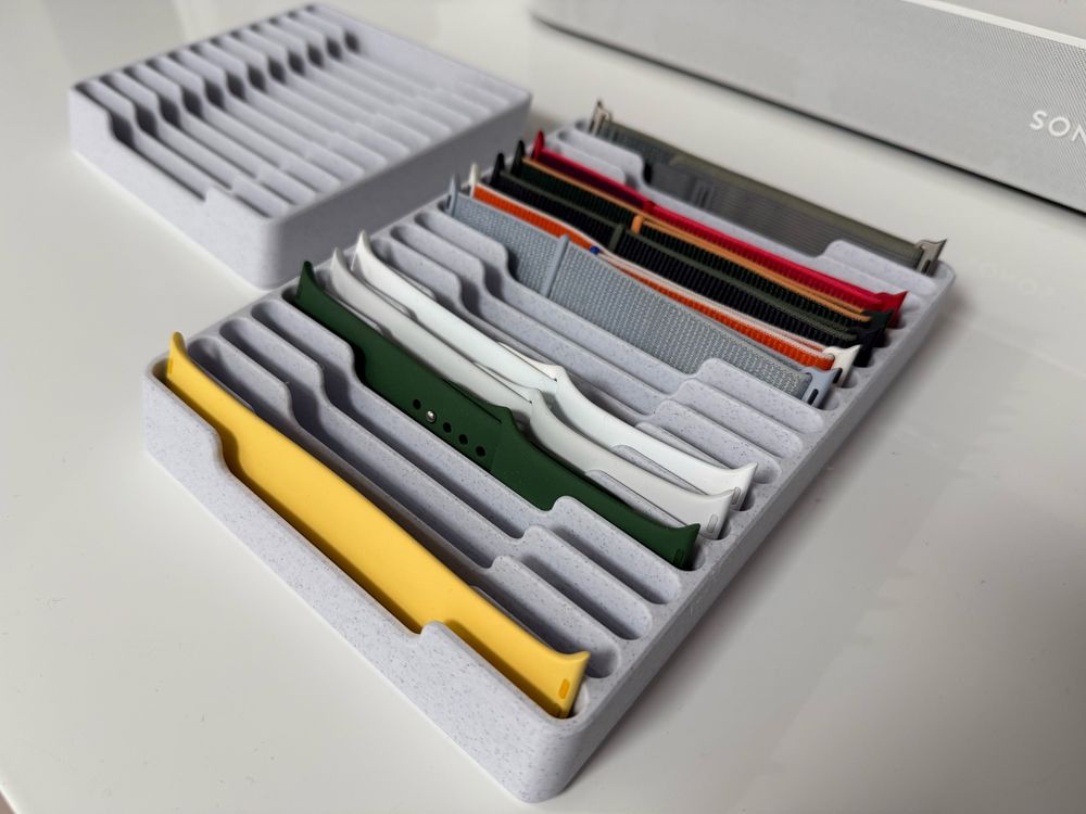 A neat, organized display of colorful watch bands stored in a gray tray with multiple compartments. The bands include silicone, fabric, and nylon materials in various colors like yellow, green, white, red, and gray, showcasing a stylish collection for interchangeable smartwatches.
