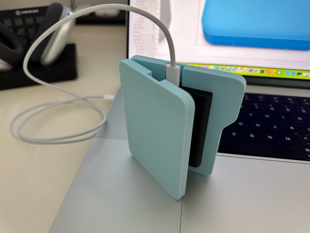 3D printed, blue Samsung SSD case in the shape of the macOS system folders. Photographed on its side on the top of a MacBoom Pro keyboard. Connected to the laptop with an USB-C cable.
