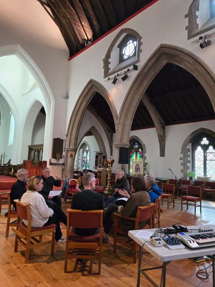 A circle of chairs with people sat talking about experimental music inside a Victorian church. Nearby is a bank of symths and other instruments. 