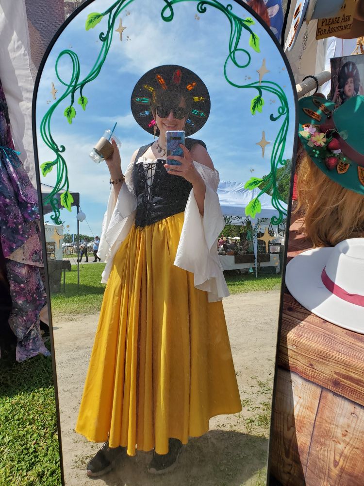 A selfie in a floor length mirror outside at the end of a booth at a fairgrounds. Siobhan is wearing a full long yellow skirt, a white chemise with long draping sleeves, a black bodice, and a black hat with stained glass acrylic windows cut into the brim. She is smiling and holding an empty cup that once held maple lemonade. The mirror has a green vine painted around the top edge.