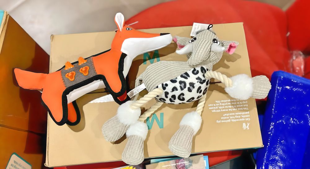 Two new dog toys on a cardboard box: one is a tough orange fox with enrichment chew nubs on flanks, and the other is a plush grey elephant with rope limbs and a leopard-print belly.