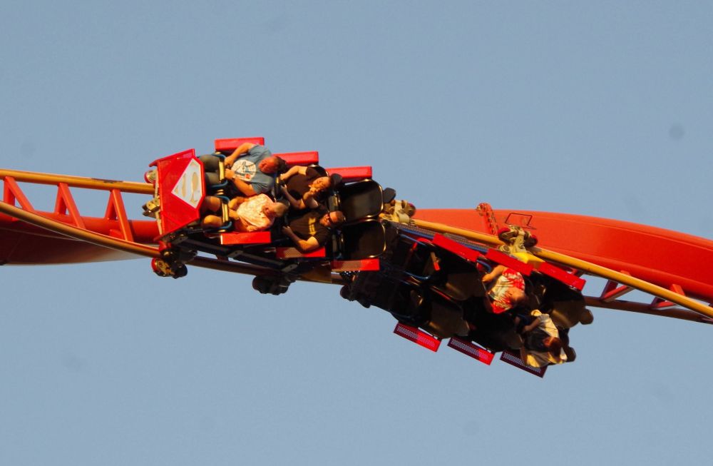 Red roller coaster train on yellow and red track.  The coaster is twisting as it comes out of an inversion so that the 2 cars of the train aren't on the same axis.  Sunset photo adding golden highlights.  Front of train has the Superman S, where the shape is distinct but S is faded to the point of being barely visible.