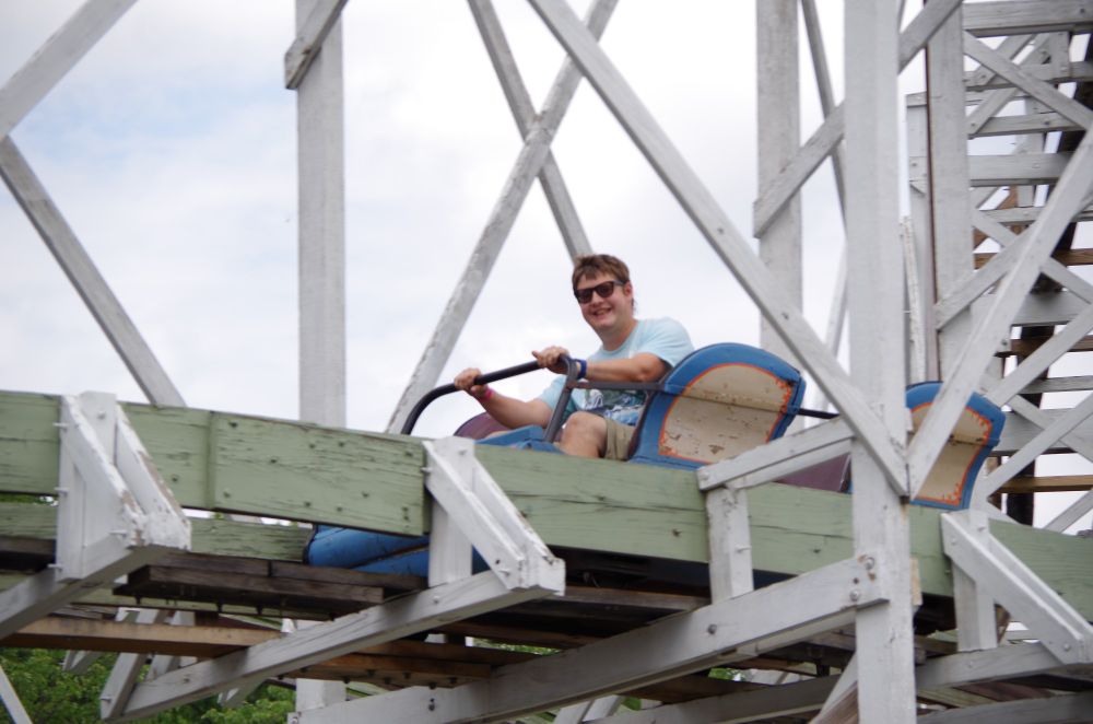 Smiling single rider on Leap the Dips roller coaster.  Small two row train surrounded by white and pale green wood track and supports.