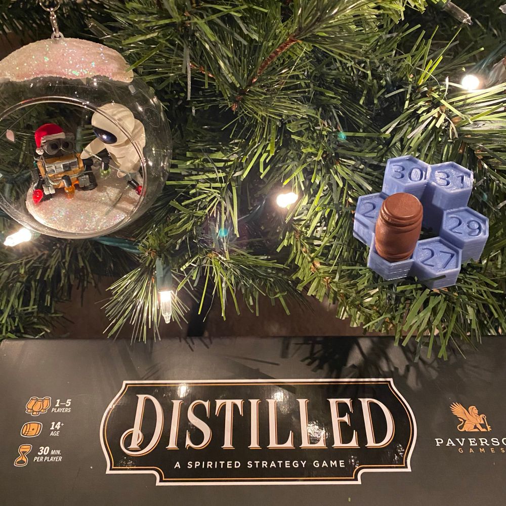 The Distilled game box spine under a Christmas tree. 

A SpielMas 3D display with a wooden barrel piece from Distilled on the 26 space rests on a lower branch. 

A Christmas ornament with WALL-E and EVE inside a glass ball helping one another untangle a sting of lights hangs from a branch to one side.