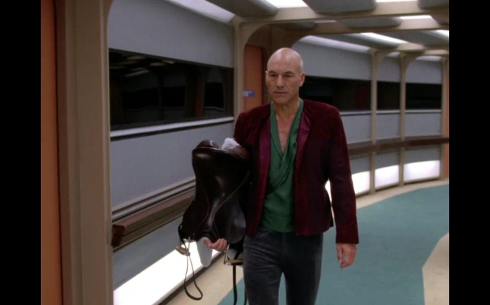 Cpt Picard in luxurious futuristic high fashion peasant garb, a red suede or satin jacket and a teal green deep v-neck blouse holds a (very new looking) saddle while walking through a corridor