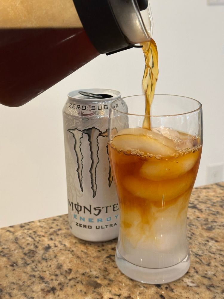 Photo of a glass with ice and Monster Zero Ultra inside and coffee is being poured over it from a Hario coffee carafe. Behind the glass is a can of Monster Zero Ultra