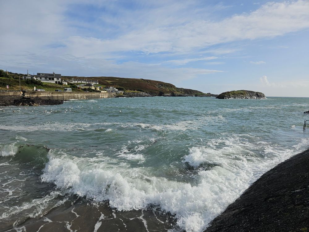 Waves crashing against a wall. On the other side of the small bay are some houses and a headland beyond. A small island is further round in the bay