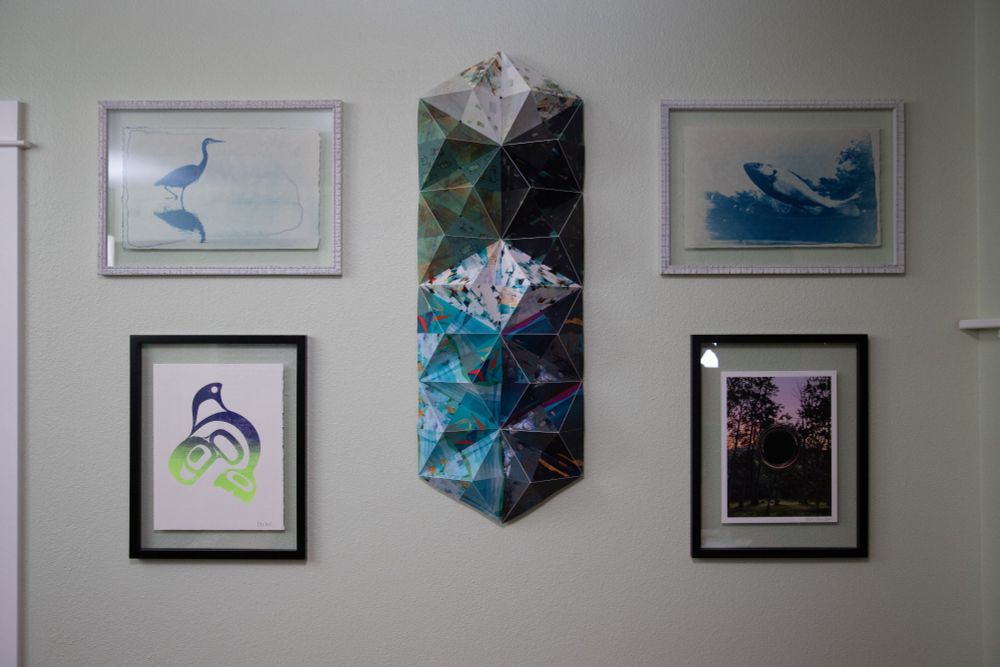 Picture of a wall with 5 works of art on the wall. In the center of the wall is a tall and narrow mixed media work consisting of multicolor prints assembled into pyramid shapes and arranged by color to look like a 3D image. The top left and top right pictures are landscape cyanotype prints of wildlife. The bottom left is an orca print and the bottom right is a surrealist painting of a forest.