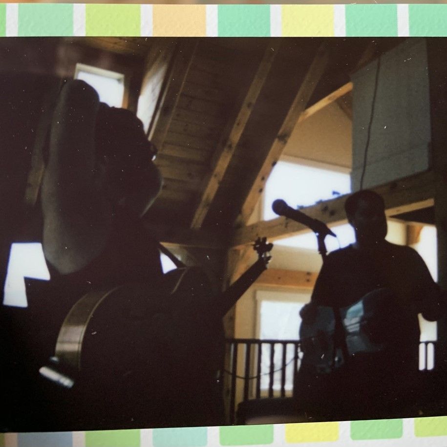 Two figures are in silhoutte, playing guitar in an old building, possibly a church. There is a green and yellow border on the edges of the photo.