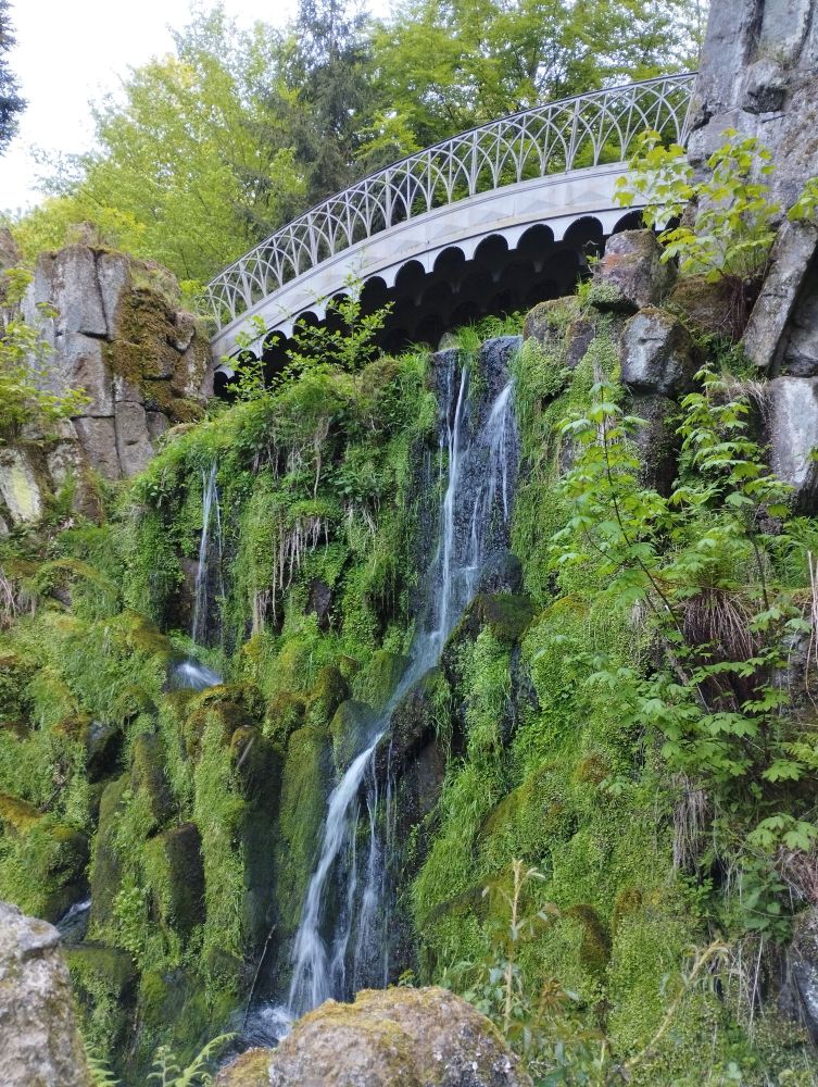 Nahaufnahme der Teufelsbrücke im Bergpark Wilhelmshöhe.
Unter der Brücke ist ein kleiner Wasserfall zu sehen. Der Abgrund ist mit Moosen und Farben zugewachsen.