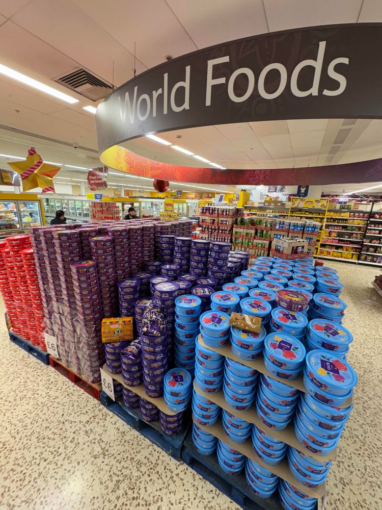 Inside a supermarket (big Tesco), World Foods is written on a massive  hoop sign dangling from the ceiling. Underneath are pallets of Celebrations, Quality Street, Miniature Heroes, and Roses chocolate selection boxes. (All very British)