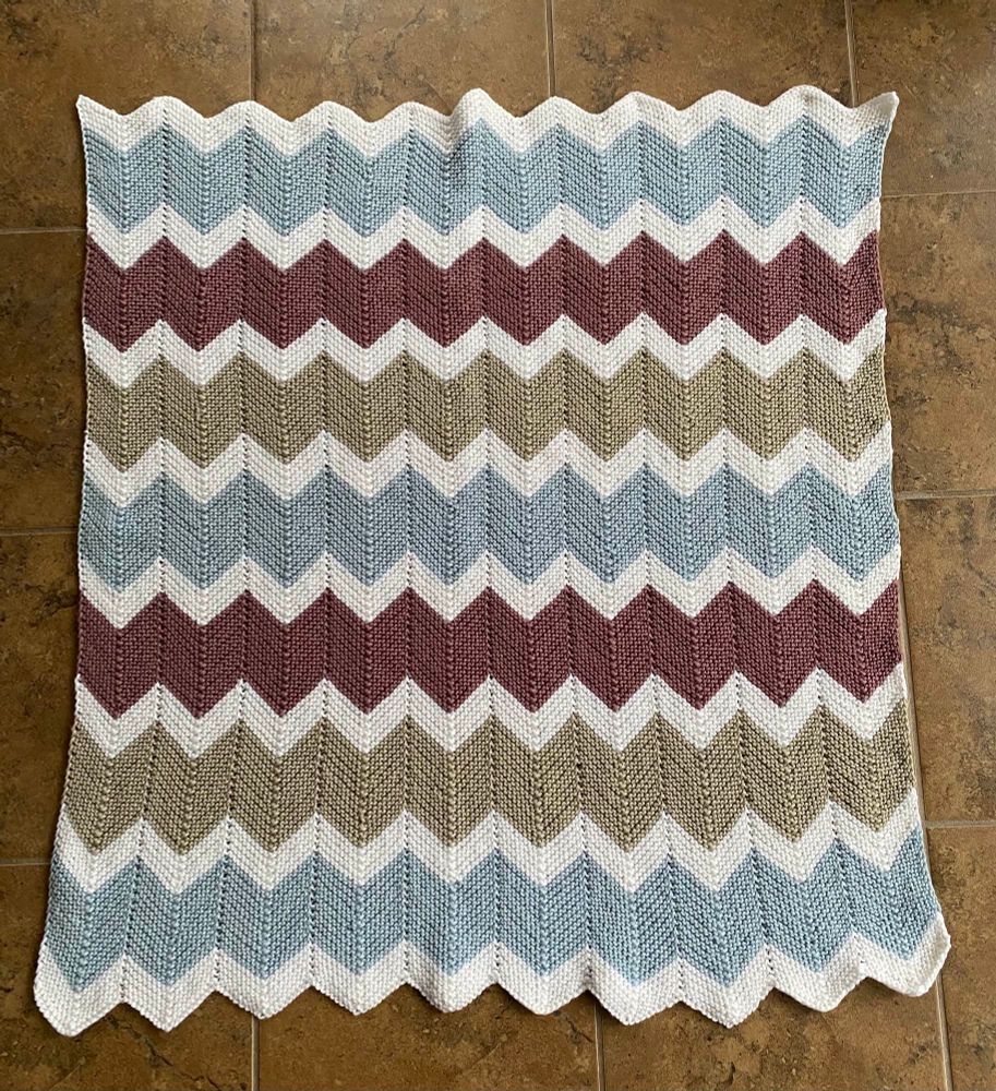 A baby blanket done in a garter stitch chevron pattern. There are smaller white chevron stripes between thicker light blue, tan, and dark mauve chevron strips. 