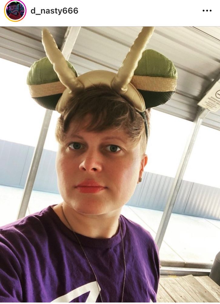 Queer wearing green ears with gold horns while standing outside wearing a purple t shirt 