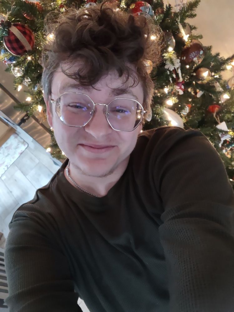 Kieran, a white transmasc with short brown hair, is standing in front of a Christmas tree and smiling. They are wearing a green long sleeve shirt and gold rimmed glasses. 